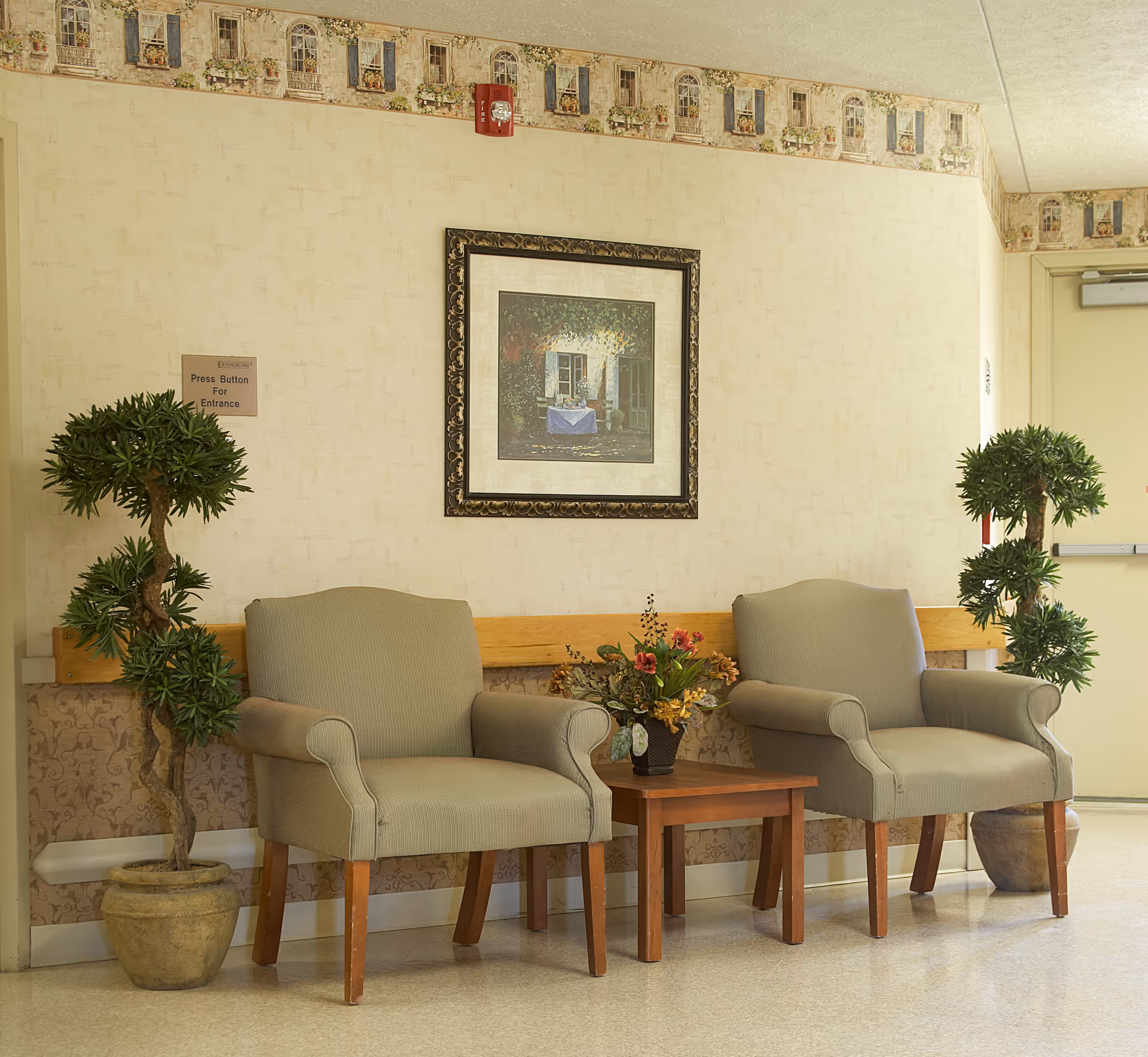 A waiting area with two beige upholstered armchairs separated by a small wooden table holding a flower arrangement. Two potted artificial trees flank the chairs. A framed painting of a table set outside a window hangs on the wall above the chairs. The wall has a wallpaper border near the ceiling and a sign that reads 'Press Button For Entrance'.