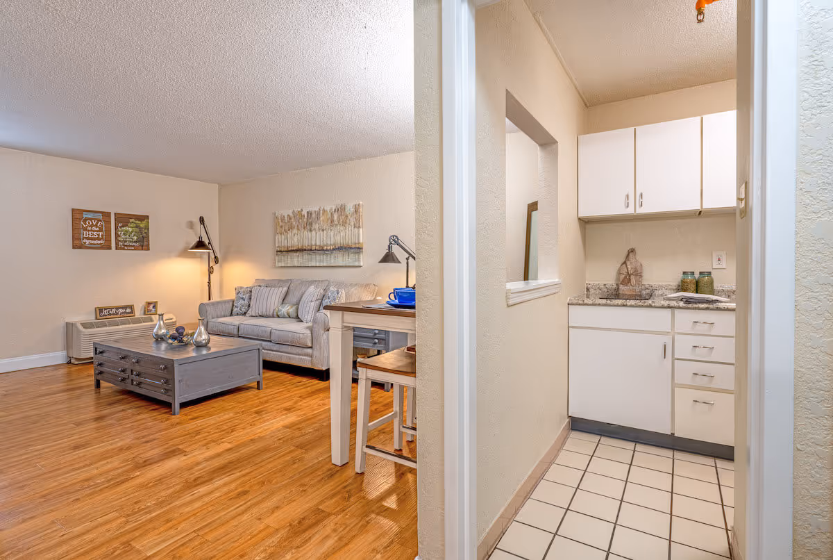 Interior view of a senior living facility showing a cozy living room with a gray sofa, a wooden coffee table, wall art, and a floor lamp. Adjacent to the living room is a small kitchen area with white cabinets, a granite countertop, and tiled flooring.