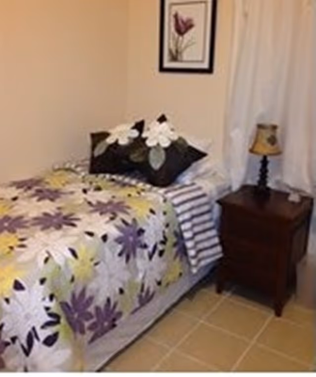 A small bedroom with a single bed covered in a floral-patterned bedspread in shades of purple, yellow, and white. There are two decorative pillows with white flowers on the bed. Next to the bed is a wooden nightstand with a small lamp and a white curtain hanging behind it. A framed picture of a flower is hung on the wall above the bed.