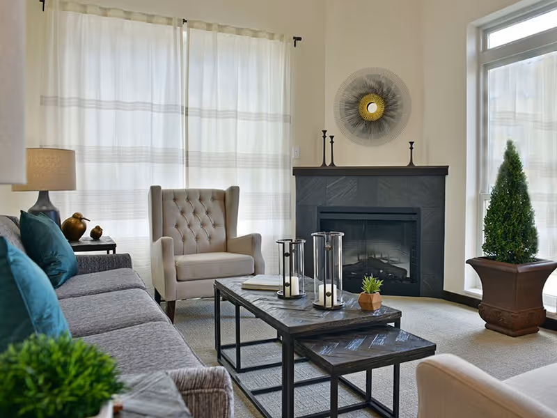 A cozy living room with a gray sofa adorned with teal pillows, a beige tufted armchair, a black fireplace with decorative items on the mantel, a coffee table with candles and a small plant, and a large window with sheer white curtains letting in natural light. There is also a potted plant near the window.