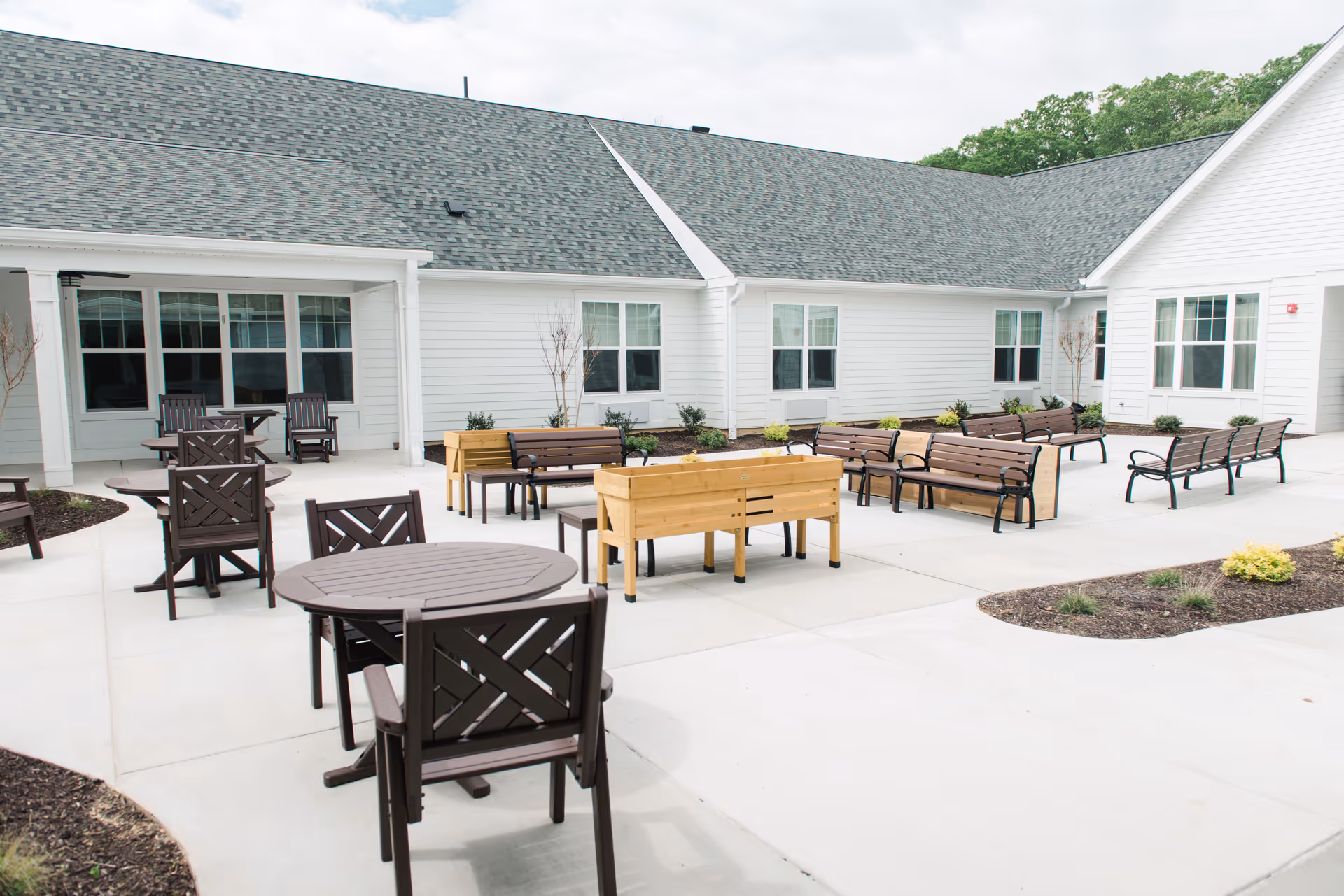Outdoor courtyard area at Legacy Ridge featuring multiple brown benches, wooden planter boxes, and round tables with chairs on a concrete patio surrounded by white buildings with gray shingled roofs and windows.