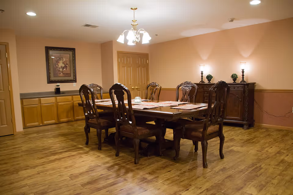 A dining room with a wooden dining table surrounded by six ornate wooden chairs. The table is set with placemats and a small centerpiece. The room has wooden flooring, beige walls, a chandelier hanging from the ceiling, and a sideboard with decorative plants and lamps.