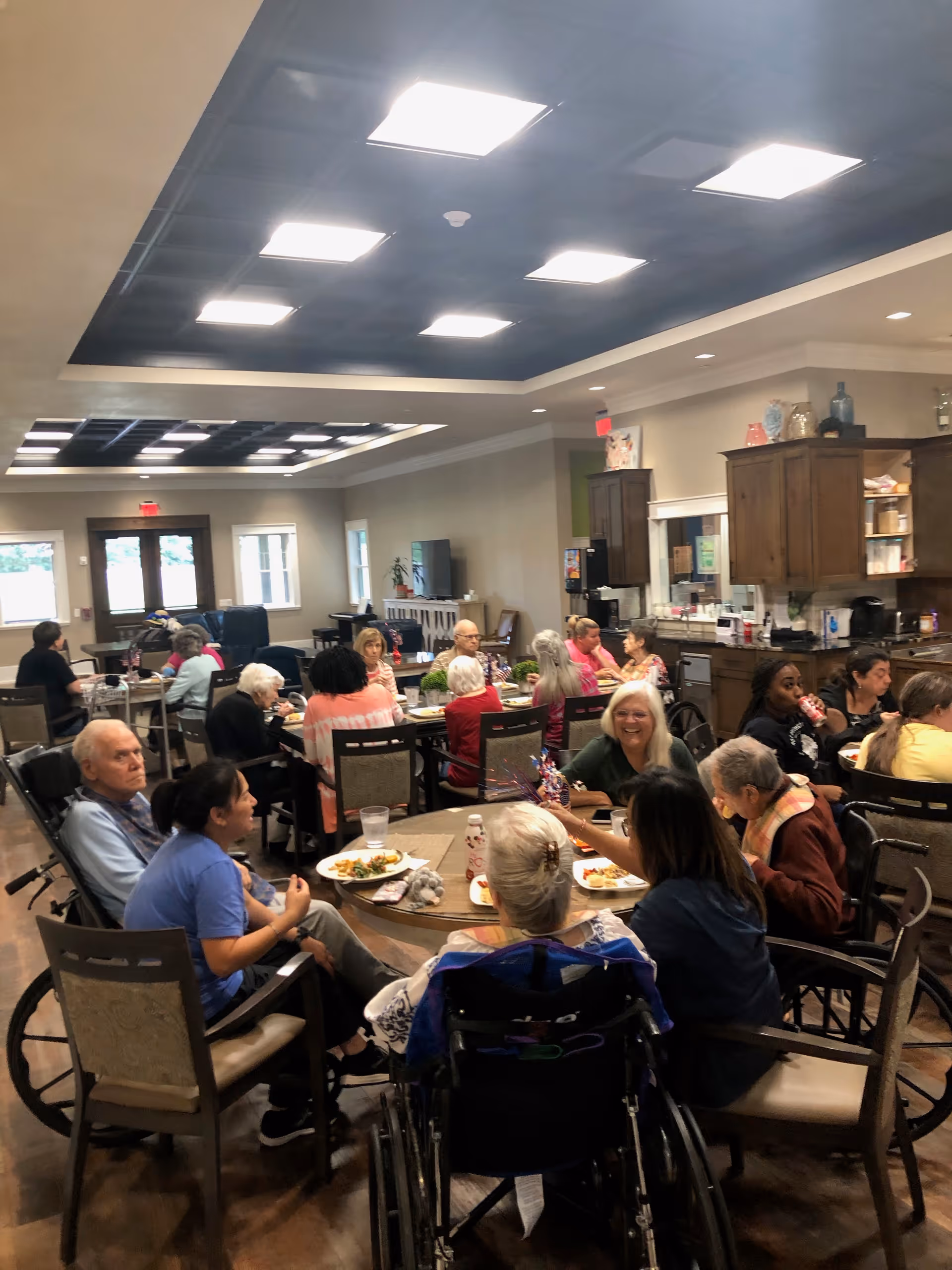 A group of elderly people and caregivers sitting around several tables in a large, well-lit dining area. Some individuals are in wheelchairs, and others are seated on chairs. The room has wooden floors, a kitchen area with cabinets on the right side, and large windows letting in natural light. People are eating, drinking, and engaging in conversation.