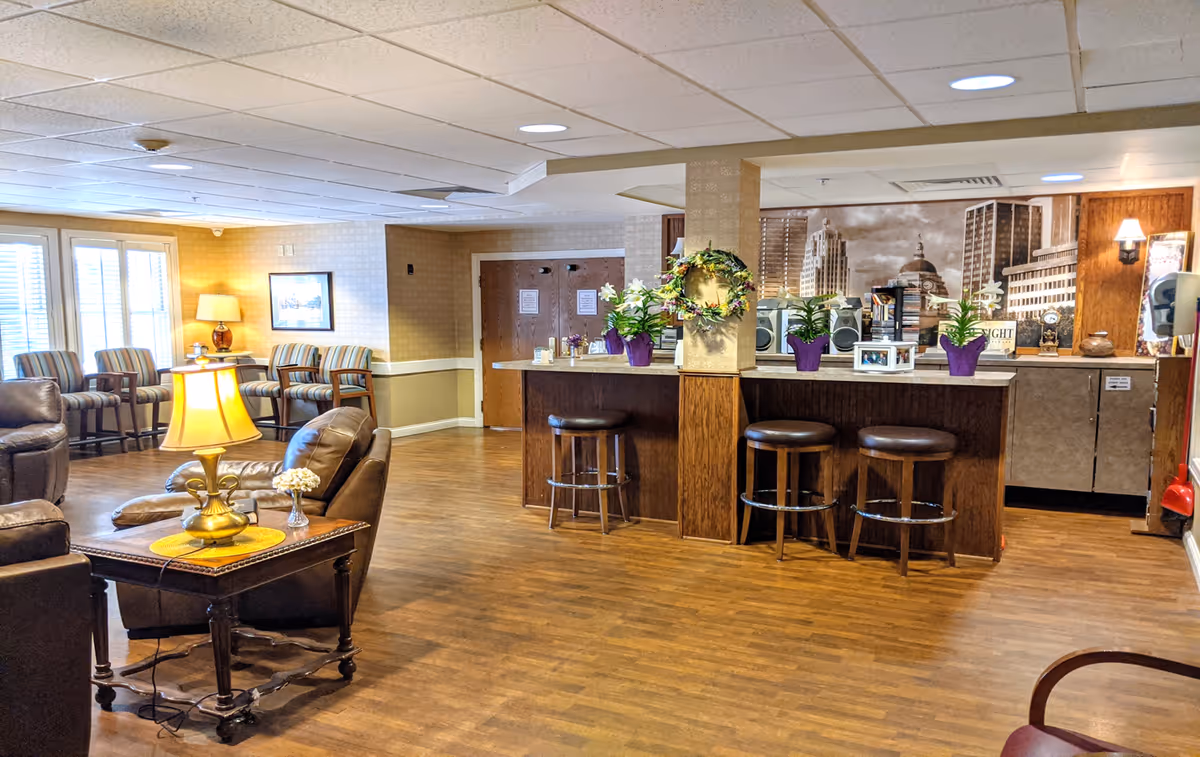 Comfortable senior living common room with leather seating, a side table lamp, striped chairs by the windows, and a counter with bar stools and potted plants.