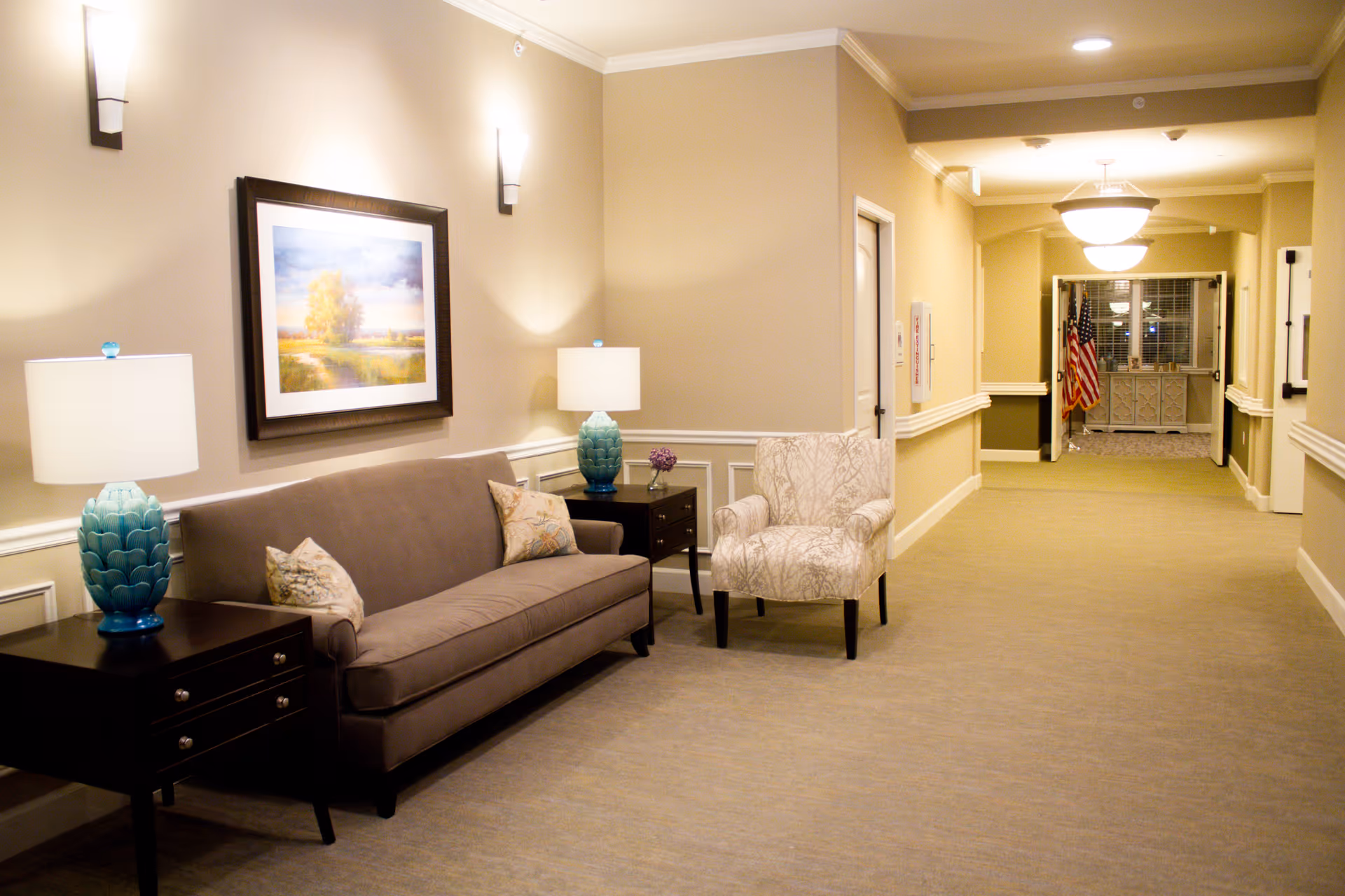A well-lit hallway in a senior living facility with beige walls and carpeted floor. The hallway features a brown sofa with two decorative pillows, a patterned armchair, two dark wooden side tables each with a blue lamp, and a framed landscape painting on the wall. At the end of the hallway, an American flag is visible near a window.