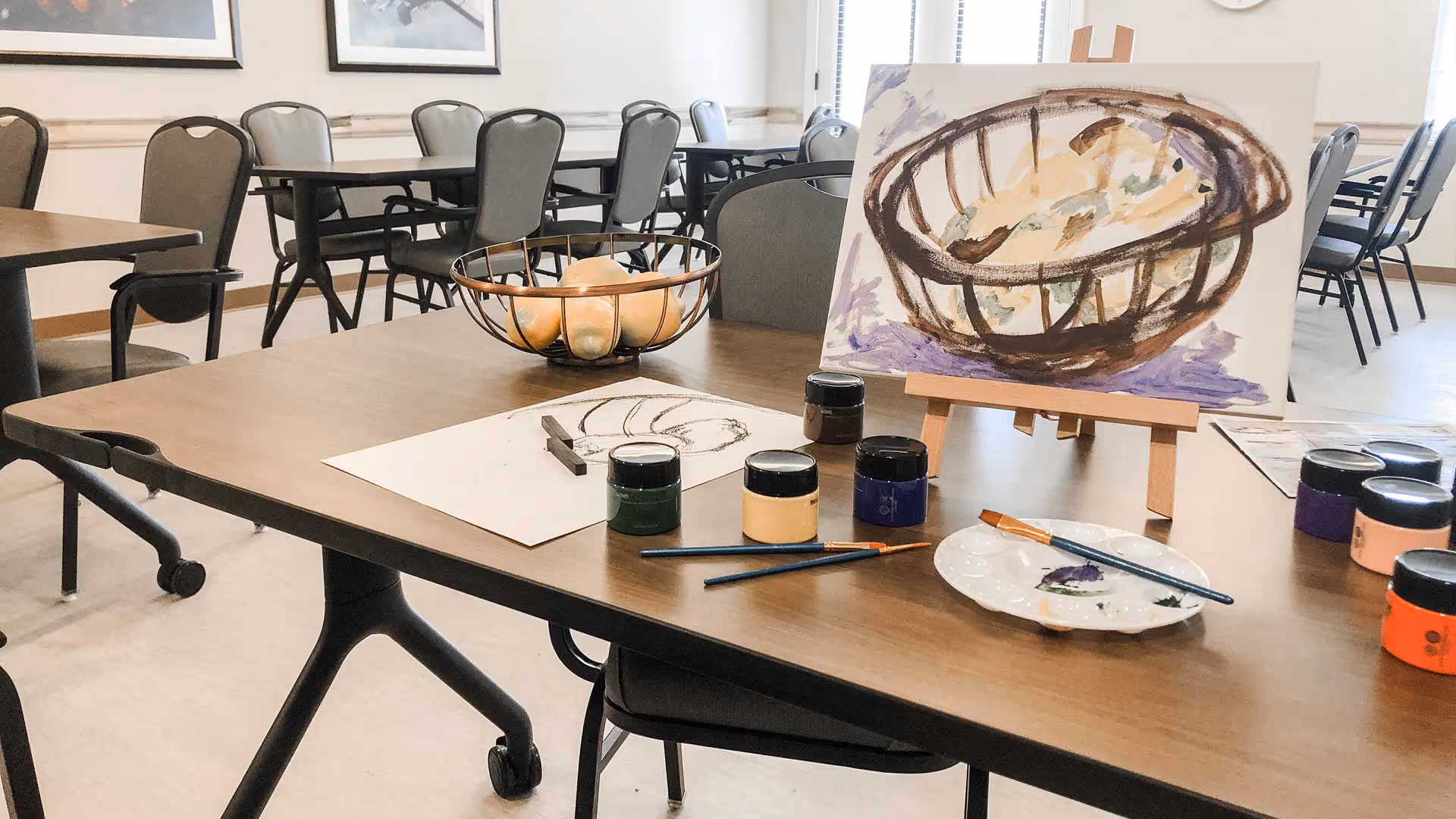A table in a communal room with painting supplies including paint jars, brushes, a palette, and a canvas on an easel showing a painting of a basket with fruit. In the background, there are multiple tables and chairs arranged in a well-lit room with framed pictures on the wall.