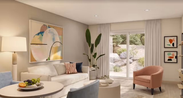 A bright and modern living room with a beige sofa adorned with decorative pillows, a round coffee table with a fruit plate, a pink armchair, and a large sliding glass door leading to an outdoor area with rocks and greenery. The room features light-colored walls, a floor lamp, abstract wall art, and a tall green plant in the corner.