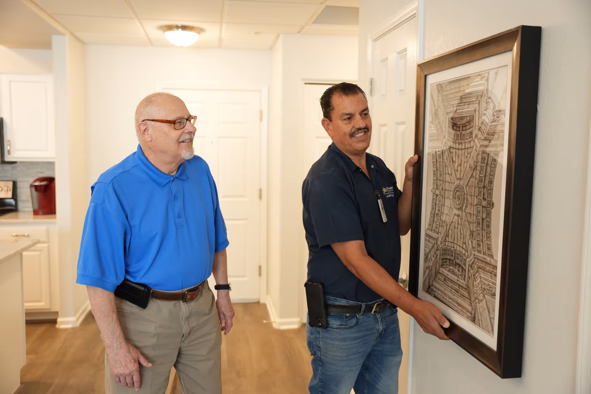 An elderly man in a blue polo shirt and khaki pants watches as a maintenance worker in a dark blue shirt and jeans hangs a framed artwork on a wall inside a well-lit room with white walls and wooden flooring.