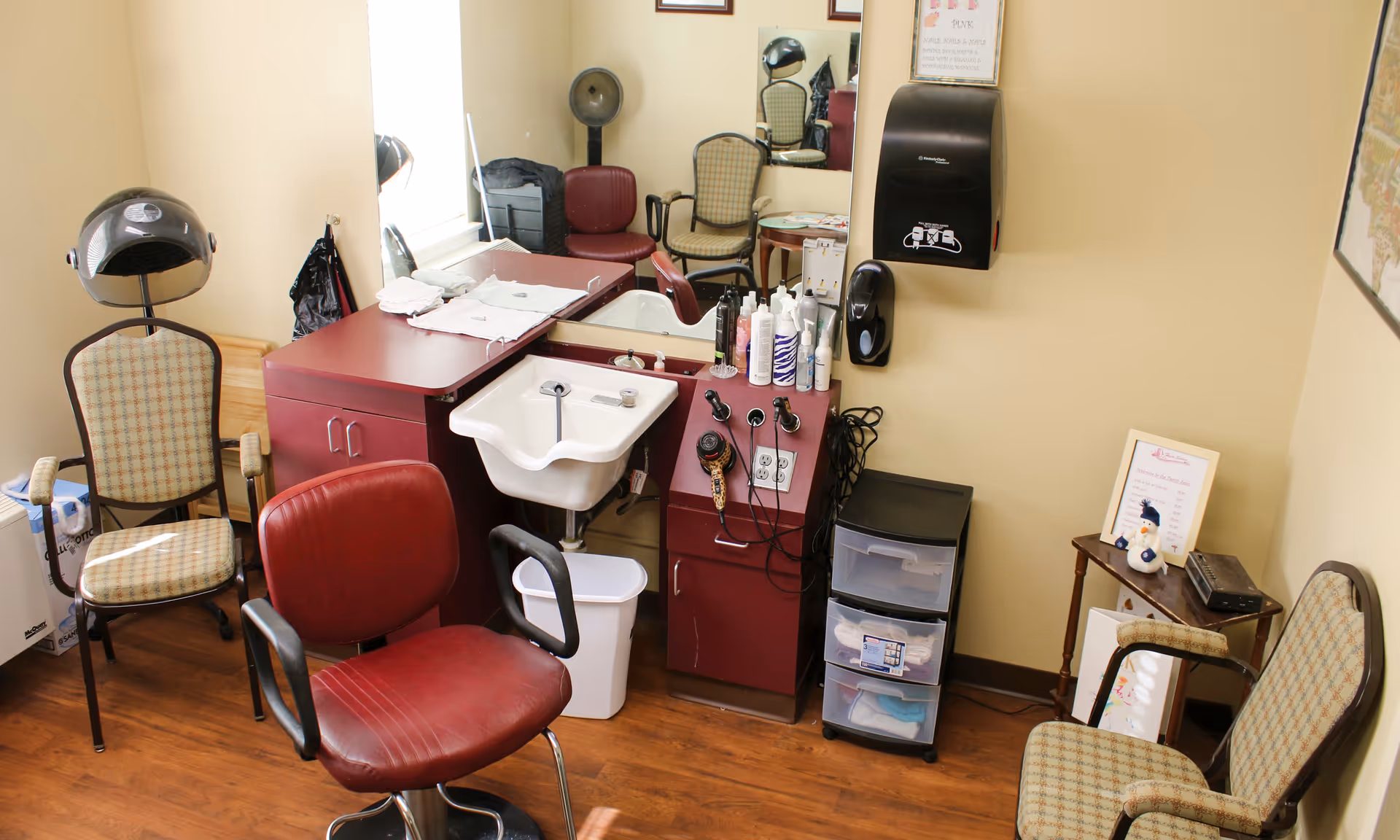 Small salon/styling room with red salon chairs, a shampoo sink, hooded hair dryer, cabinets and styling supplies.