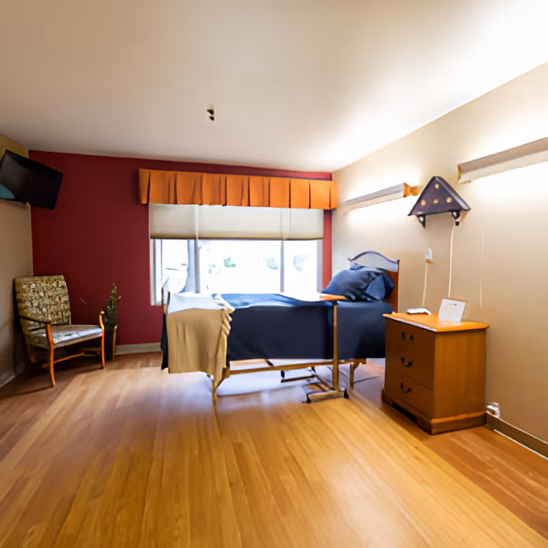 A single patient bedroom with a hospital-style bed, nightstand, armchair, and a large window with an orange valance.