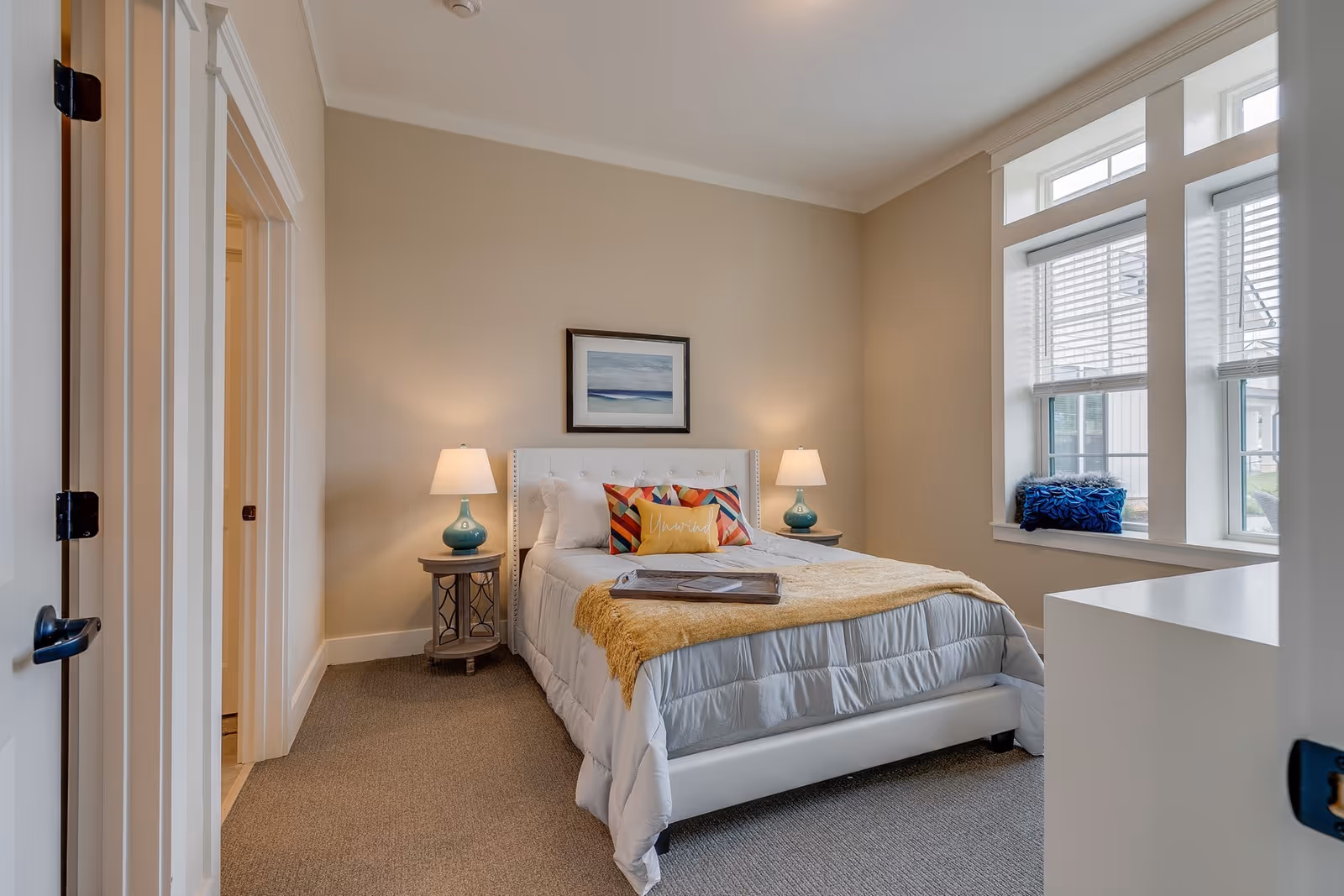 Bright furnished bedroom with a bed, colorful pillows and throw, two bedside lamps, and a large window with blinds.