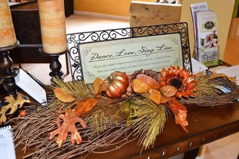 A decorative autumn-themed centerpiece on a wooden table featuring artificial leaves, a small pumpkin, and a sunflower. Behind the centerpiece is a framed sign that reads 'Dance, Love, Sing, Live' along with some brochures and two large candles on candle holders.