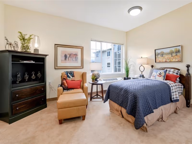 A cozy bedroom in a senior living facility featuring a neatly made bed with a blue quilt and decorative pillows, a tan armchair with a red cushion, a small round side table with a lamp and plant, a black cabinet with decorative items, and two framed paintings on the walls. A large window lets in natural light.