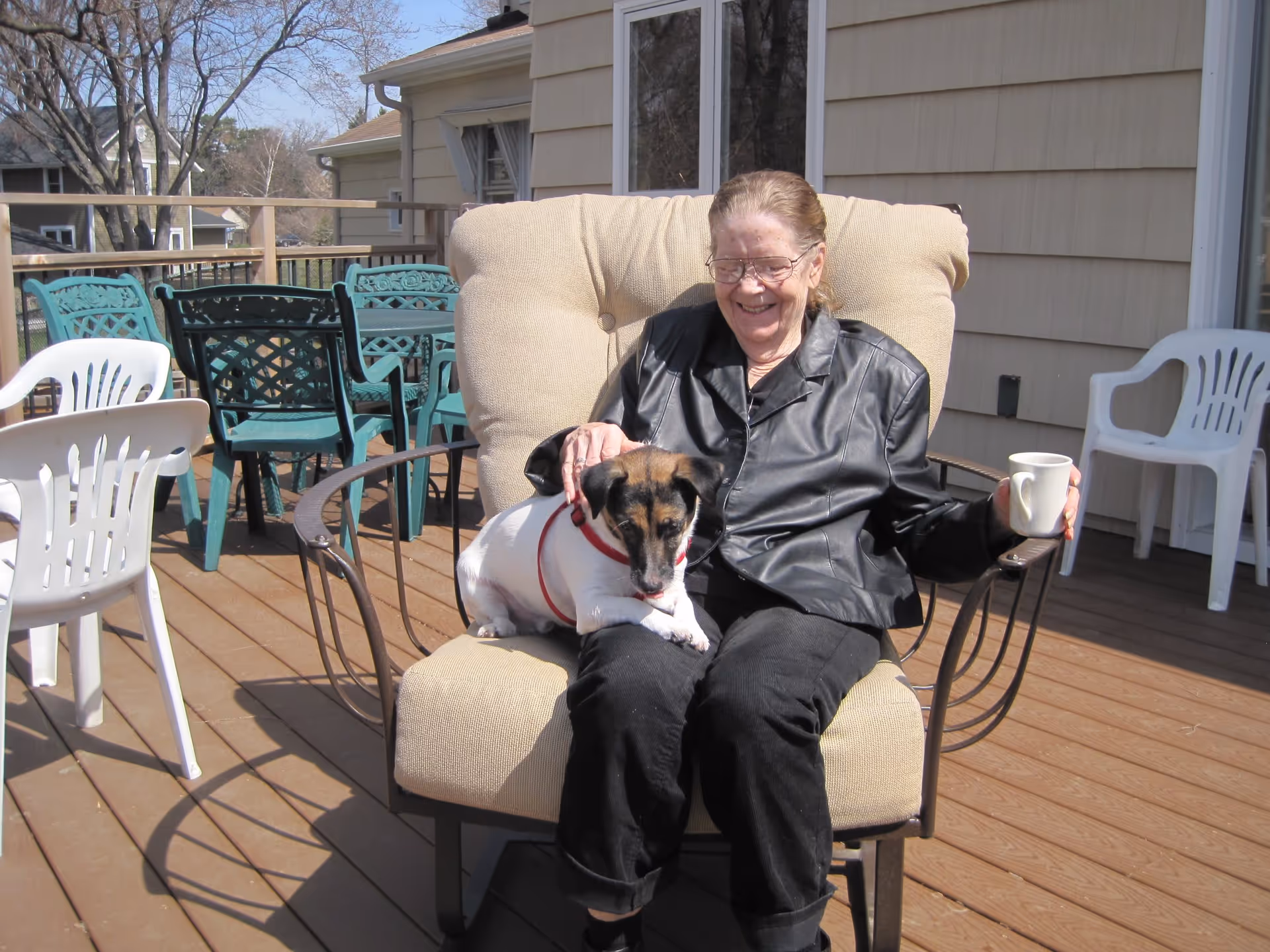 An elderly woman wearing glasses and a black leather jacket sits on a cushioned outdoor chair on a wooden deck, smiling while holding a white mug in one hand and petting a small white and brown dog sitting on her lap. The deck has several plastic chairs and a table, with houses and leafless trees visible in the background.