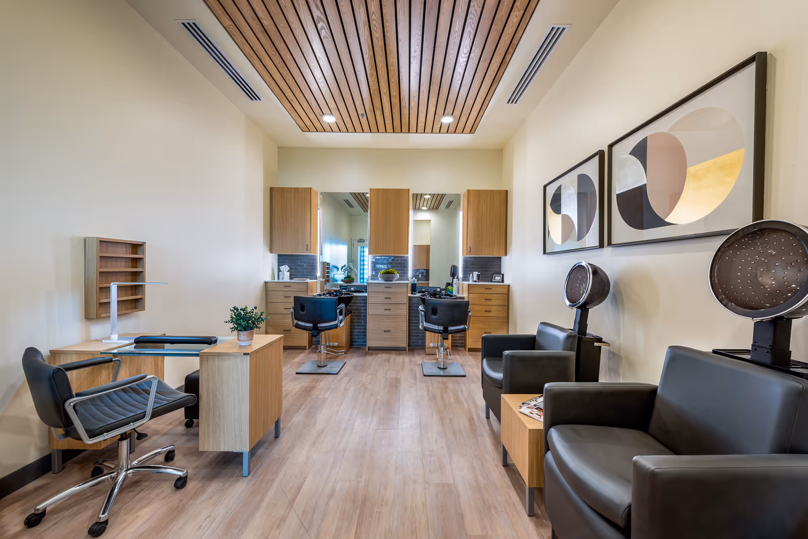 Interior view of a modern salon area with two black salon chairs in front of mirrors and wooden cabinets. To the right, there are two black leather chairs with hair dryers above them and a small wooden side table with magazines. On the left, there is a small desk with a black chair and a potted plant. The ceiling has wooden slats with recessed lighting, and two abstract framed artworks hang on the right wall.