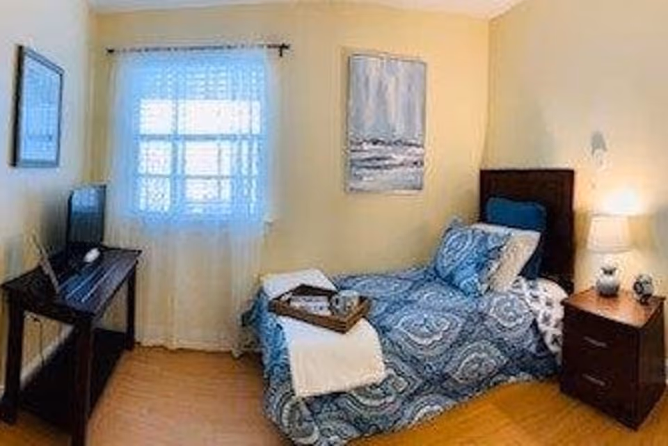 Small cozy bedroom with a single bed in blue patterned bedding, a nightstand with a lamp, a window with sheer curtains, and a TV on a console.