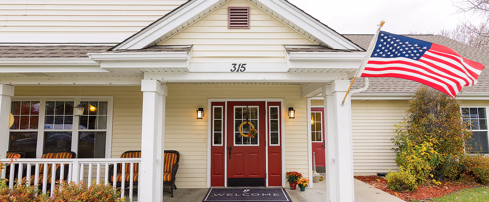 Front entrance of a building with beige siding and a red double door. There is a porch with white railings and several striped cushioned chairs. An American flag is mounted on the right side of the porch. The number 315 is displayed above the door, and a welcome mat is placed in front of the entrance. Some bushes and plants are visible around the porch area.