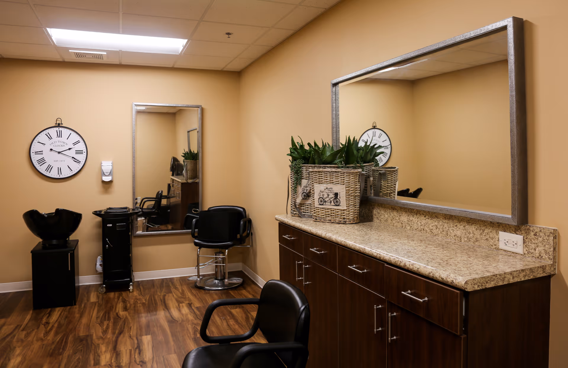 Interior view of a hair salon area with beige walls and wood flooring. There are two black salon chairs, a black hair washing station, a wall clock with Roman numerals, a large rectangular mirror above a granite countertop with dark wooden cabinets, and a decorative basket with green plants on the counter.