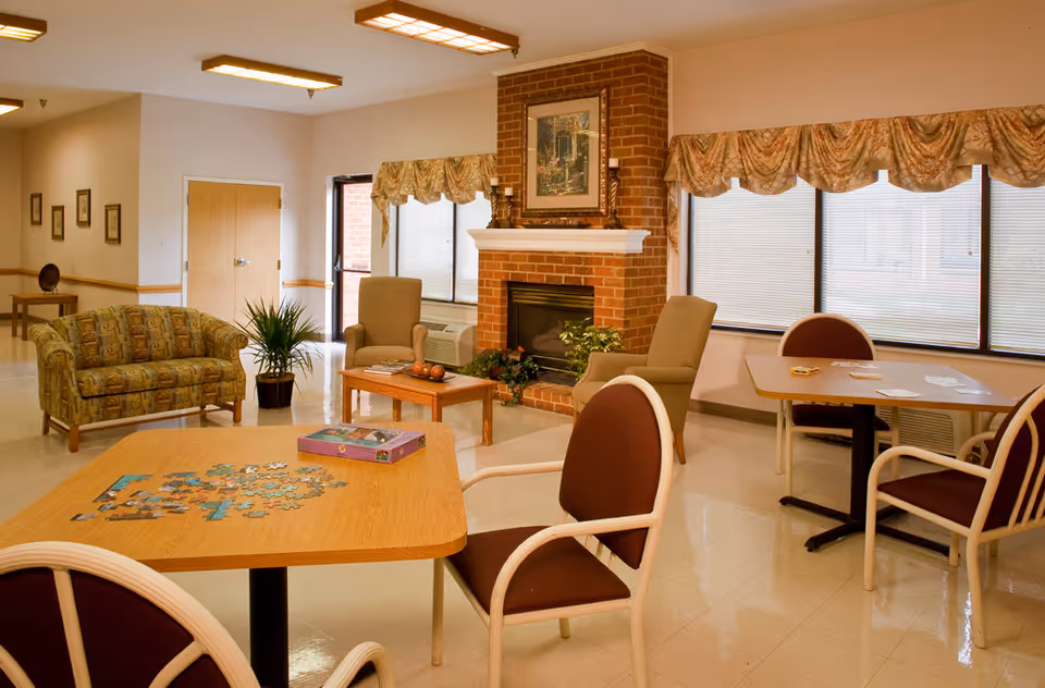 A cozy common area in a senior living facility featuring a brick fireplace with a framed picture above it, two armchairs, a patterned loveseat, and two tables with chairs. One table has a partially completed jigsaw puzzle and the other has playing cards. Large windows with valances let in natural light, and there are plants and framed pictures on the walls.