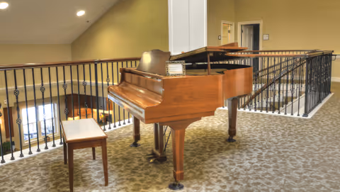 A wooden grand piano with an open lid and a matching bench is placed on a carpeted area near a black metal railing overlooking a lower level. The space has beige walls and ceiling lights, with doors visible in the background.