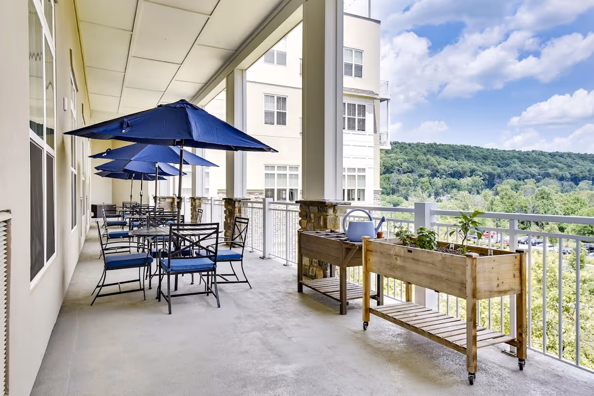 Covered outdoor balcony with tables, blue umbrellas, chairs and planter boxes overlooking tree-covered hills.