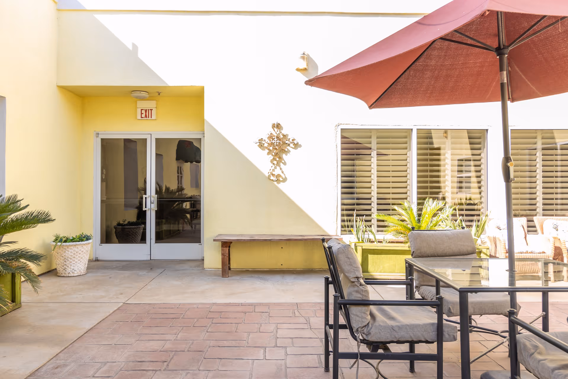 Outdoor patio area with a glass table and cushioned chairs under a large red umbrella. There are potted plants and a bench against a yellow wall with a decorative metal cross. Double glass doors with an exit sign above them are visible in the background.