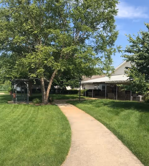 A paved walkway curves through a well-maintained grassy area with large leafy trees providing shade. To the right, there is a covered patio area attached to a building with outdoor seating. The sky is clear and blue.