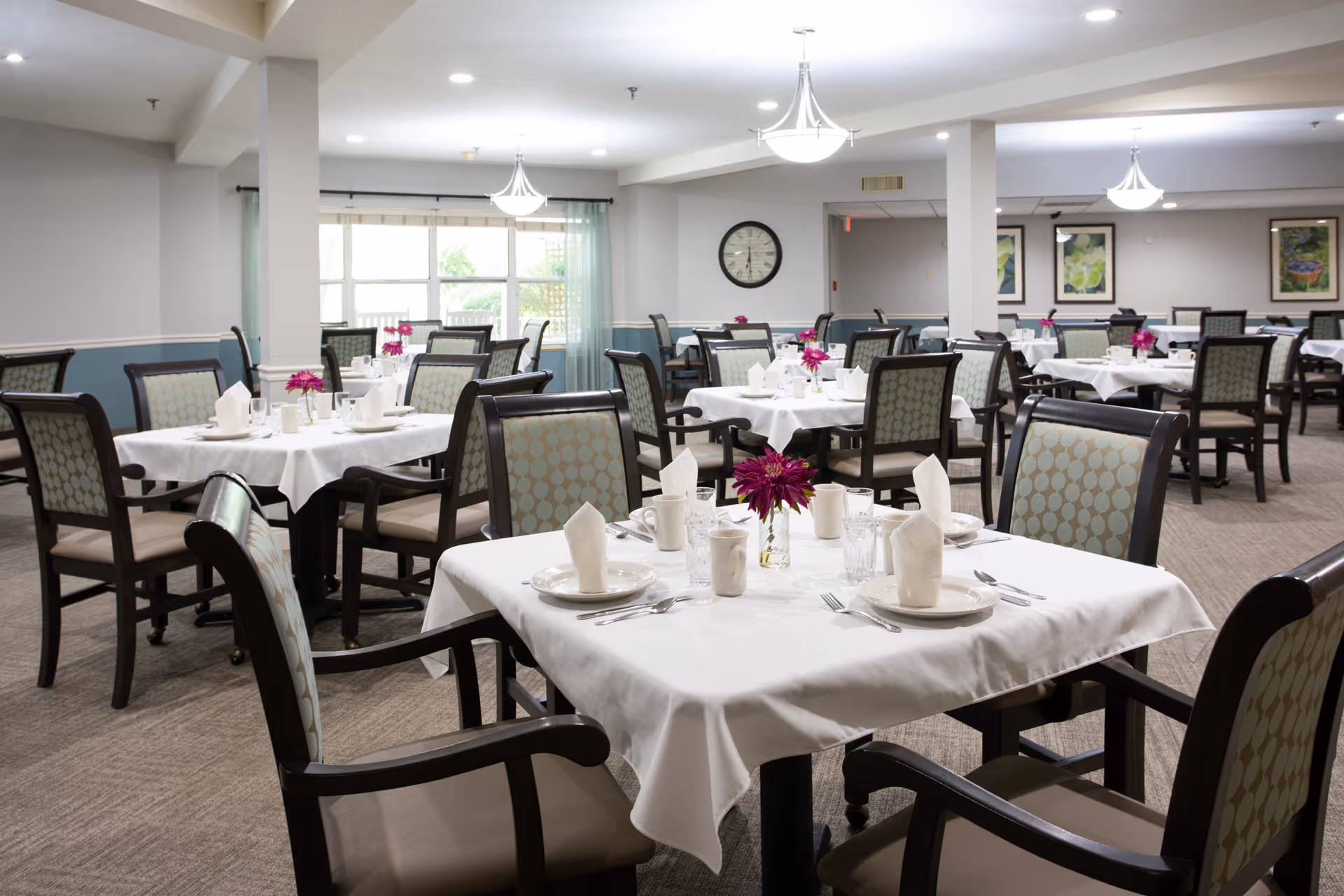 Bright dining room with multiple tables set with white tablecloths, napkins, tableware, and small floral centerpieces.