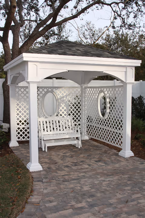 White wooden gazebo with a shingled roof and lattice walls on two sides, featuring two oval mirrors and a white wooden bench swing inside, situated on a paved stone patio with trees and greenery in the background.