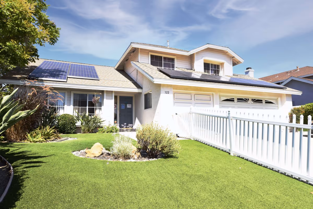 Front exterior view of a two-story house with solar panels on the roof, a well-maintained green lawn, various plants and shrubs, a white picket fence, and a clear blue sky.