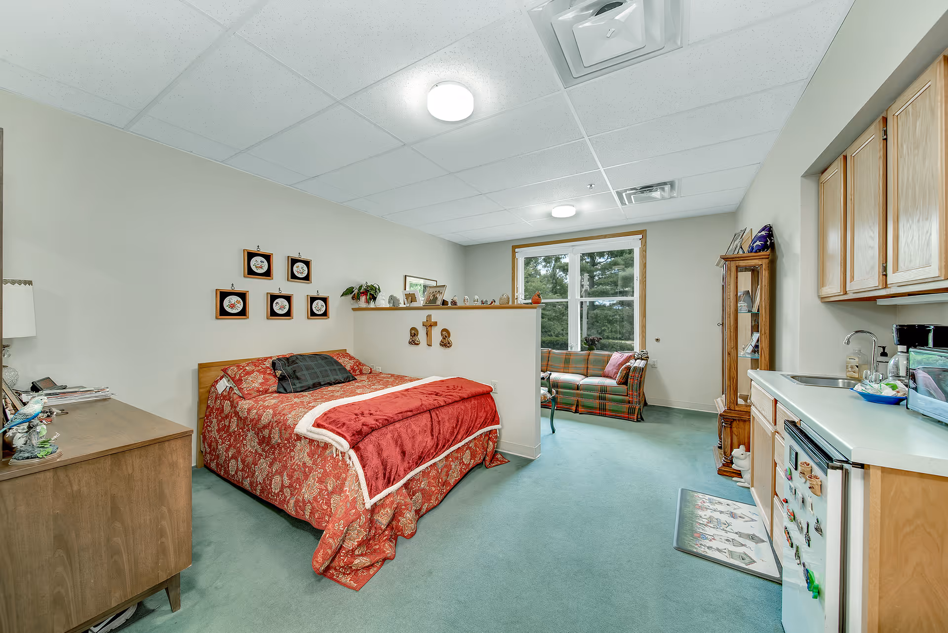 A senior living facility room featuring a bed with red patterned bedding, a wooden dresser, a small kitchenette with a sink and mini fridge, a plaid sofa near a large window, and various decorative items on shelves and walls.