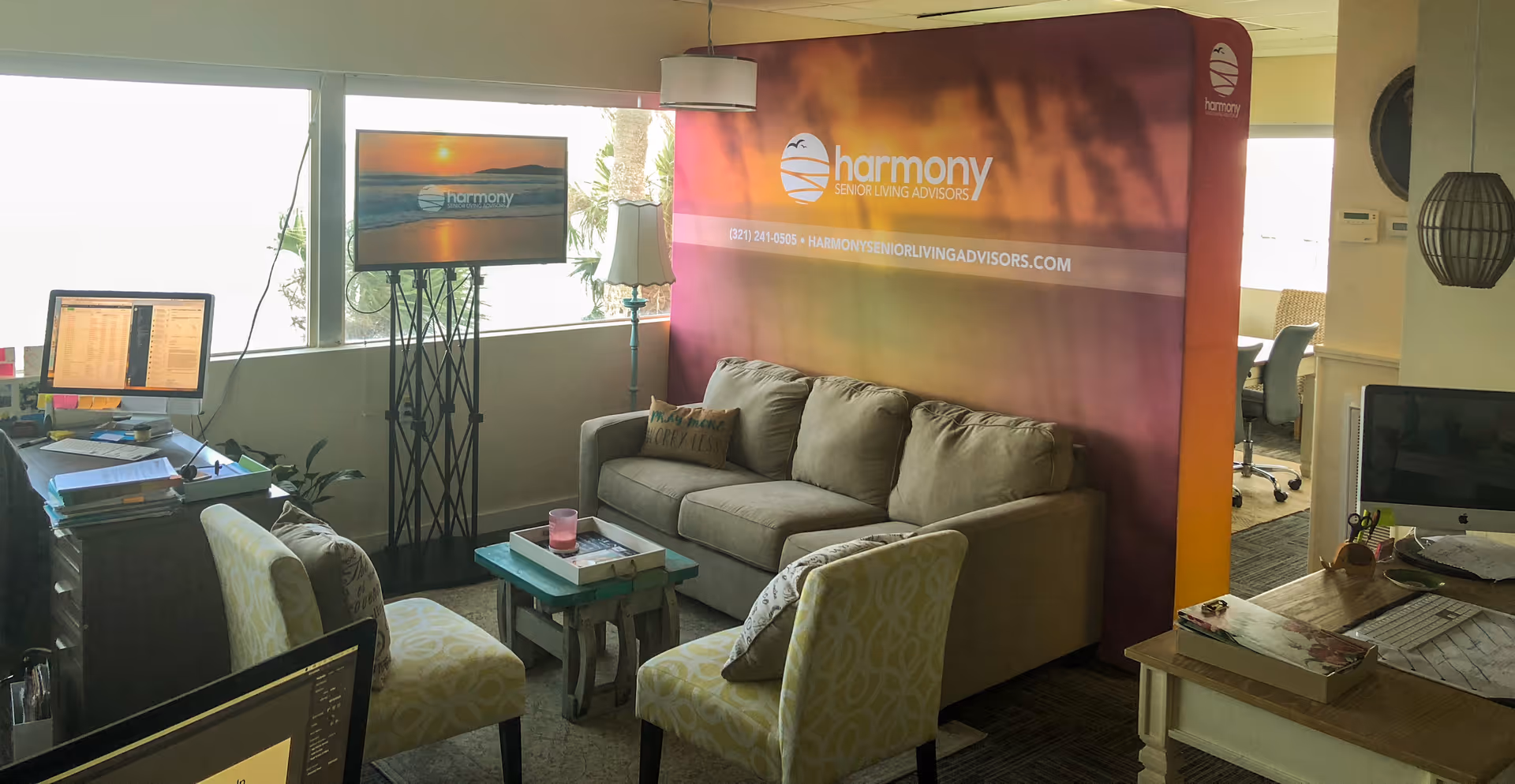 An office space with a beige couch, two patterned chairs, a small coffee table, and multiple computer desks with monitors. A large banner with the Harmony Senior Living Advisors logo and contact information is displayed behind the couch. A TV screen on a stand also shows the Harmony logo. The room has large windows letting in natural light.