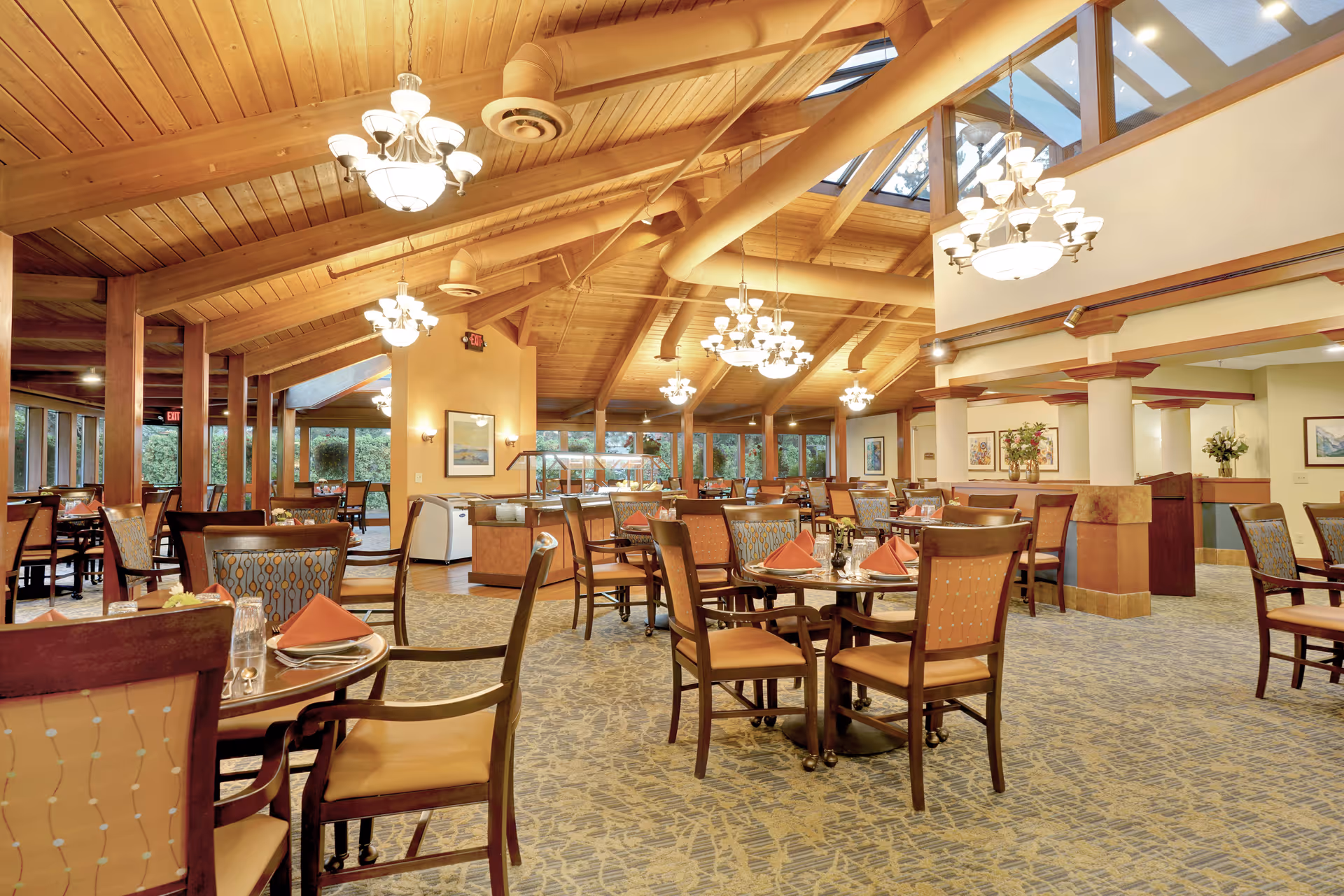 Spacious dining room with wooden vaulted ceiling and multiple chandeliers. Several round tables are set with chairs, napkins, and tableware. Large windows along the walls provide natural light and a view of greenery outside. The room has a warm and inviting atmosphere with carpeted floors and decorative wall art.