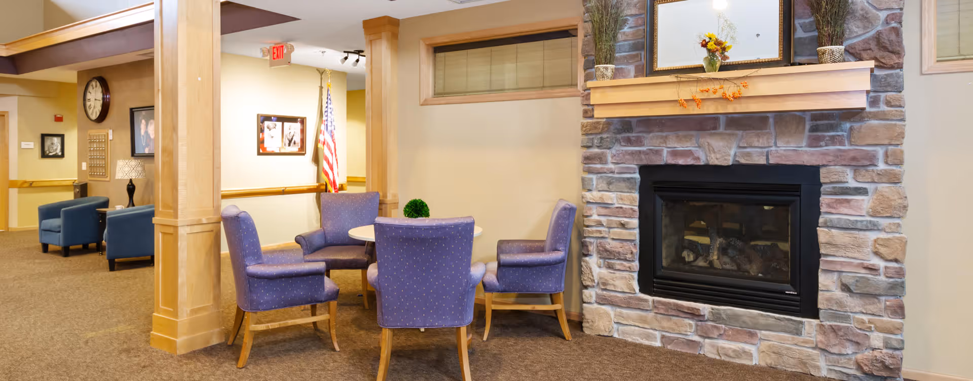 A cozy seating area in an assisted living facility featuring four purple upholstered chairs arranged around a small round table. To the right, there is a stone fireplace with a wooden mantel decorated with a small plant and a framed mirror. The background shows a hallway with additional seating and an American flag near the wall.