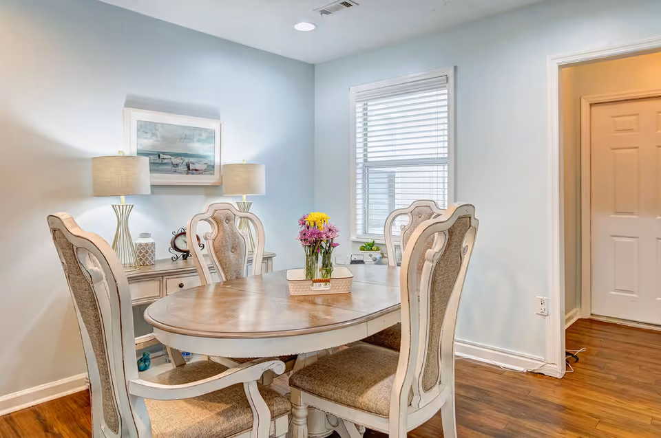 A dining room with a wooden oval table surrounded by four upholstered chairs. On the table is a small tray with a vase of colorful flowers. Behind the table is a sideboard with two table lamps and decorative items, and a framed painting of a beach scene hangs on the wall. A window with white blinds lets in natural light, and the floor is wooden.
