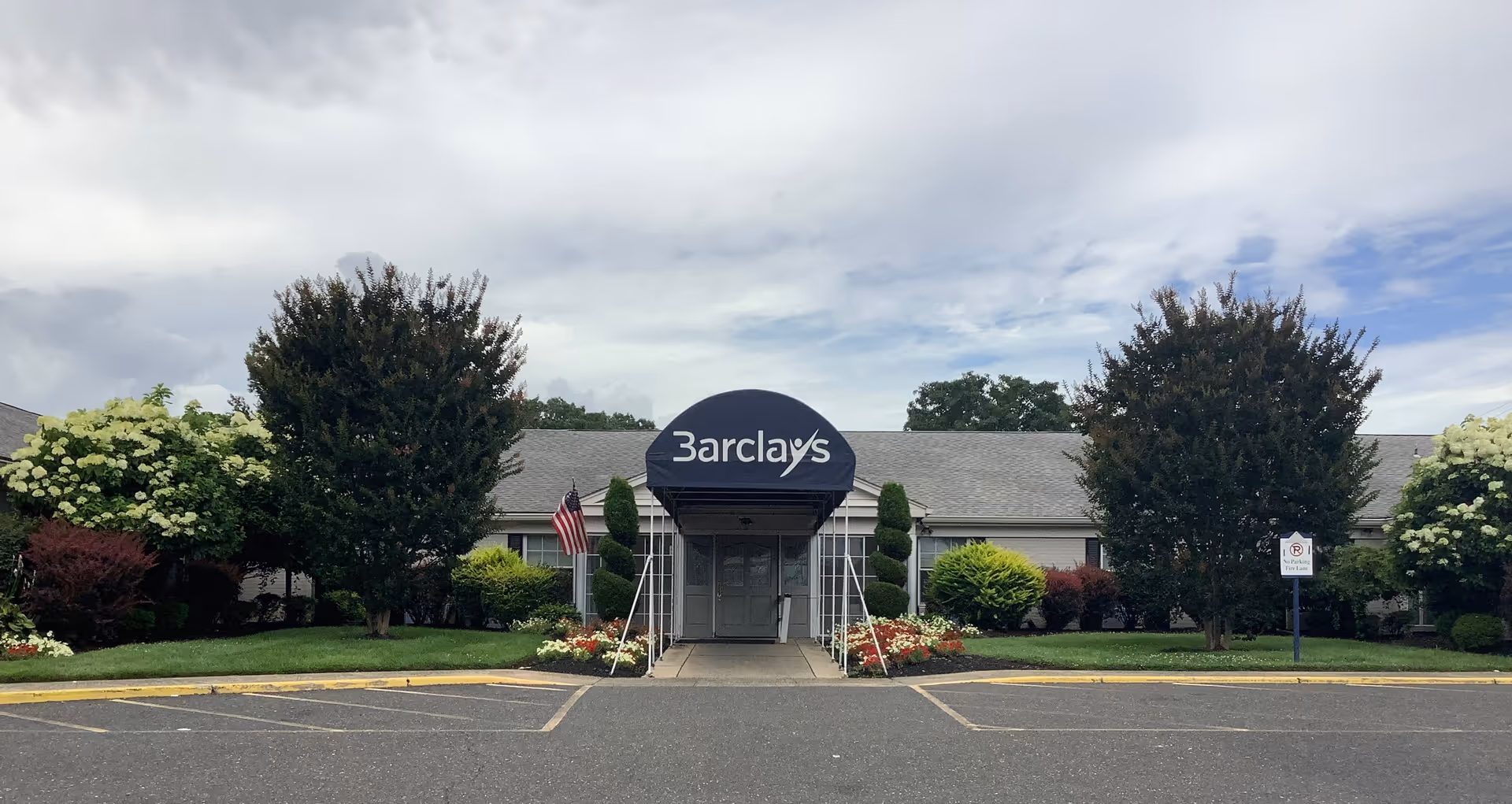 Front exterior view of Barclays Rehabilitation And Health Care Center with a covered entrance, surrounded by well-maintained landscaping including trees, bushes, and flowers under a partly cloudy sky.