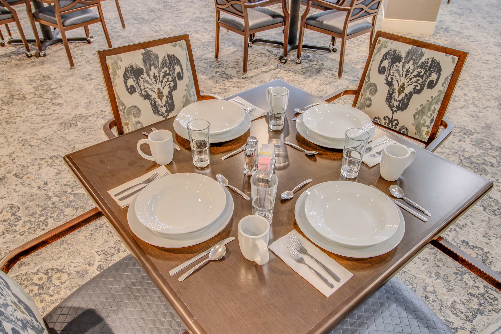 A square wooden dining table set for four with white plates, bowls, mugs, glasses, silverware, and napkins. The chairs around the table have patterned upholstery with a floral design. The background shows more tables and chairs on a carpeted floor.