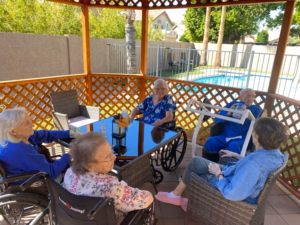 Five elderly individuals sitting and socializing under a wooden gazebo with lattice walls. Three of them are in wheelchairs, and two are seated in wicker chairs around a glass-top table. In the background, there is a fenced swimming pool and greenery.