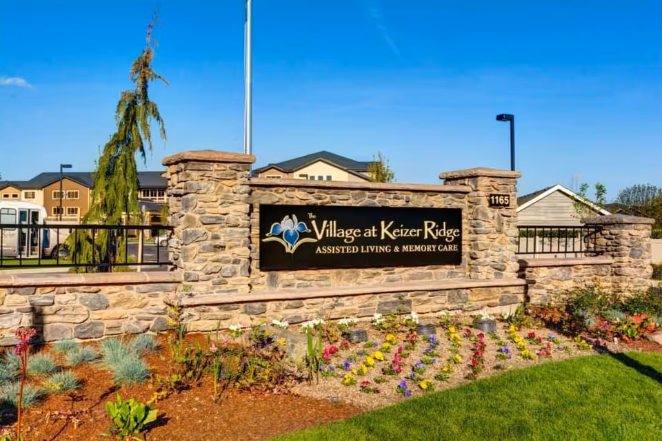 Stone entrance sign for The Village at Keizer Ridge assisted living and memory care with landscaped flowers and facility buildings behind it.