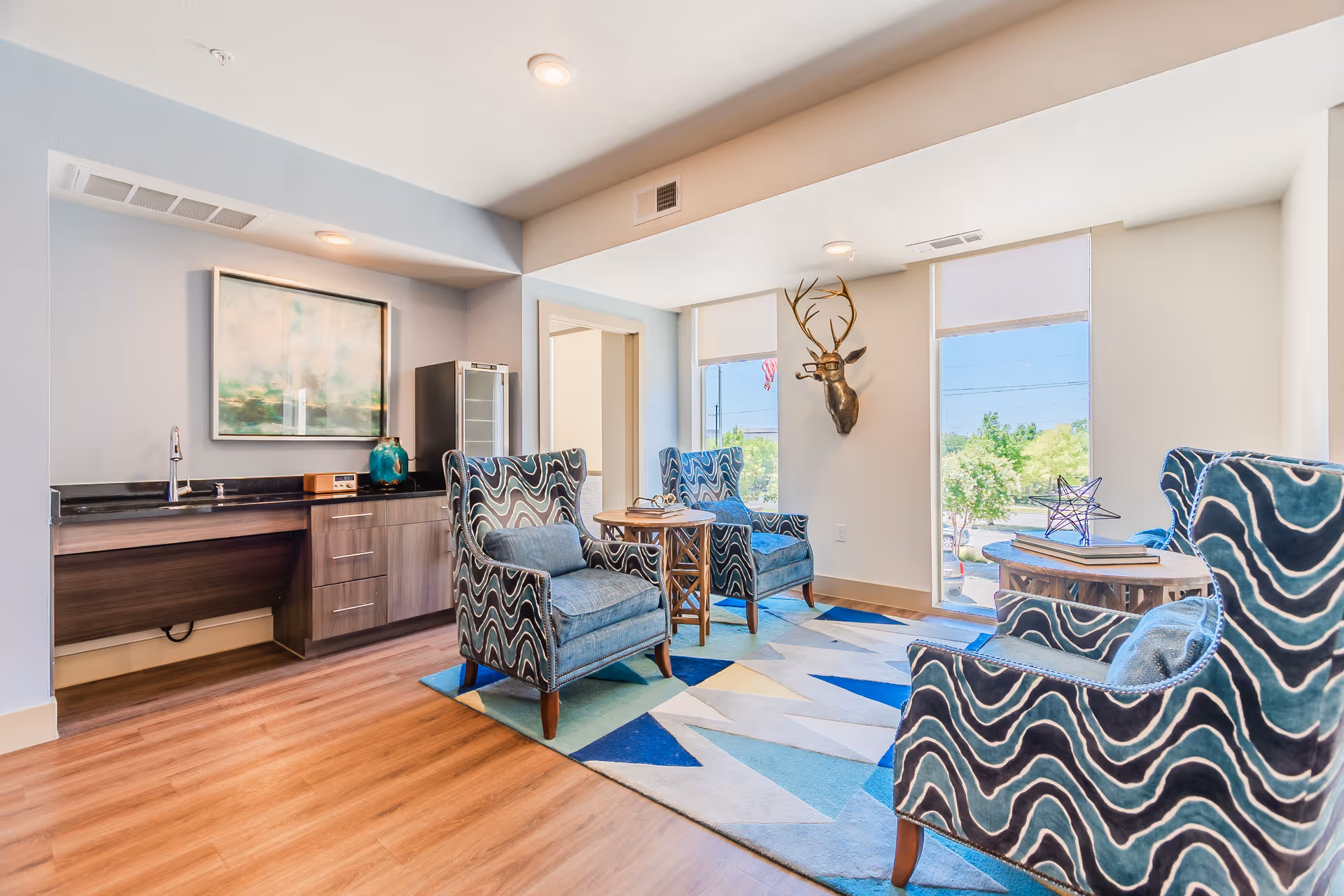 A bright and modern sitting area with four patterned blue armchairs arranged around two wooden side tables on a geometric patterned rug. The room features large windows with white roller shades, a mounted decorative deer head sculpture on the wall, and a small kitchenette area with a sink, cabinets, a mini fridge, and a framed abstract painting above the counter.