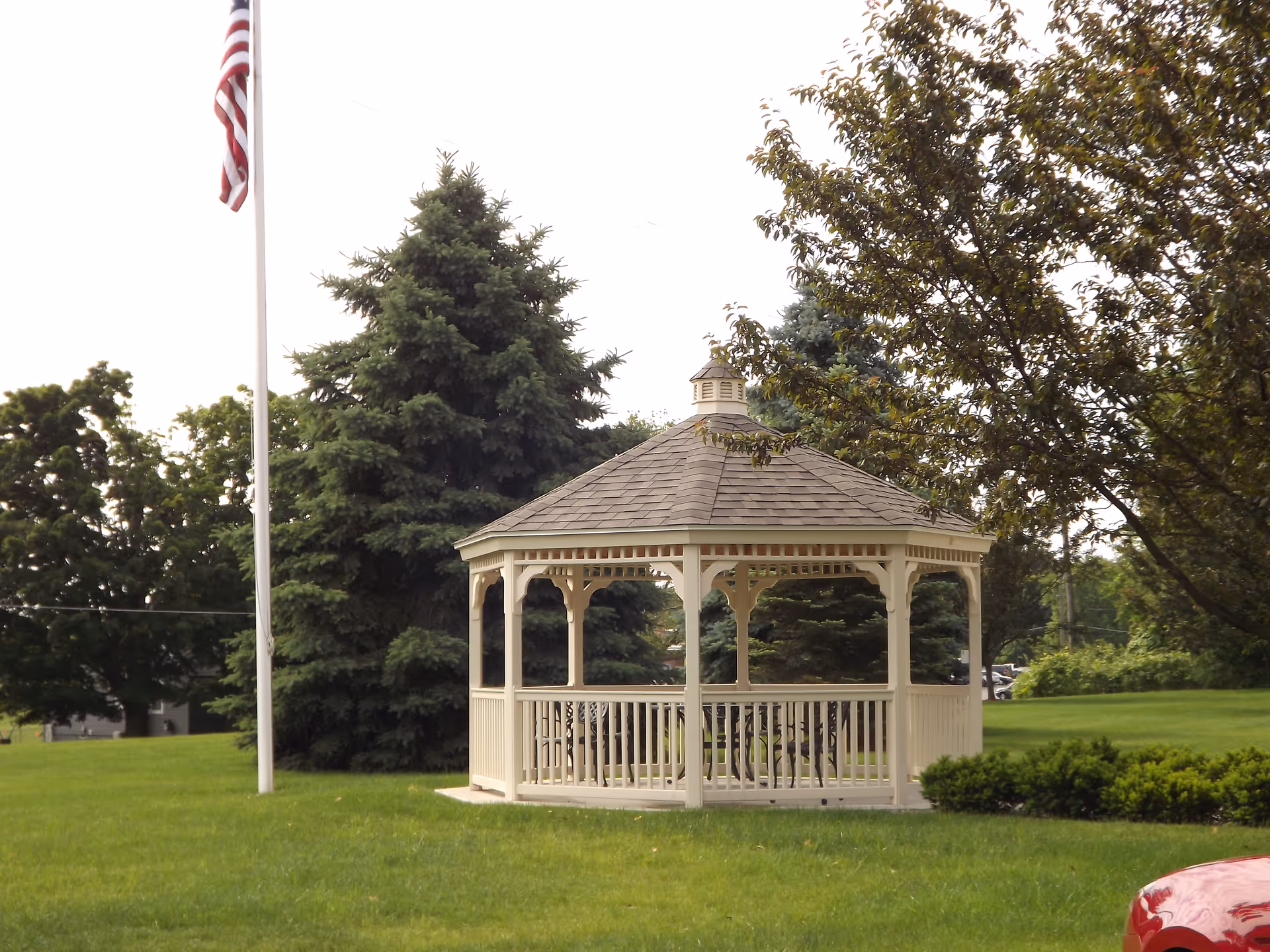 A white wooden gazebo with a shingled roof situated on a green lawn surrounded by trees, with an American flag on a flagpole nearby.
