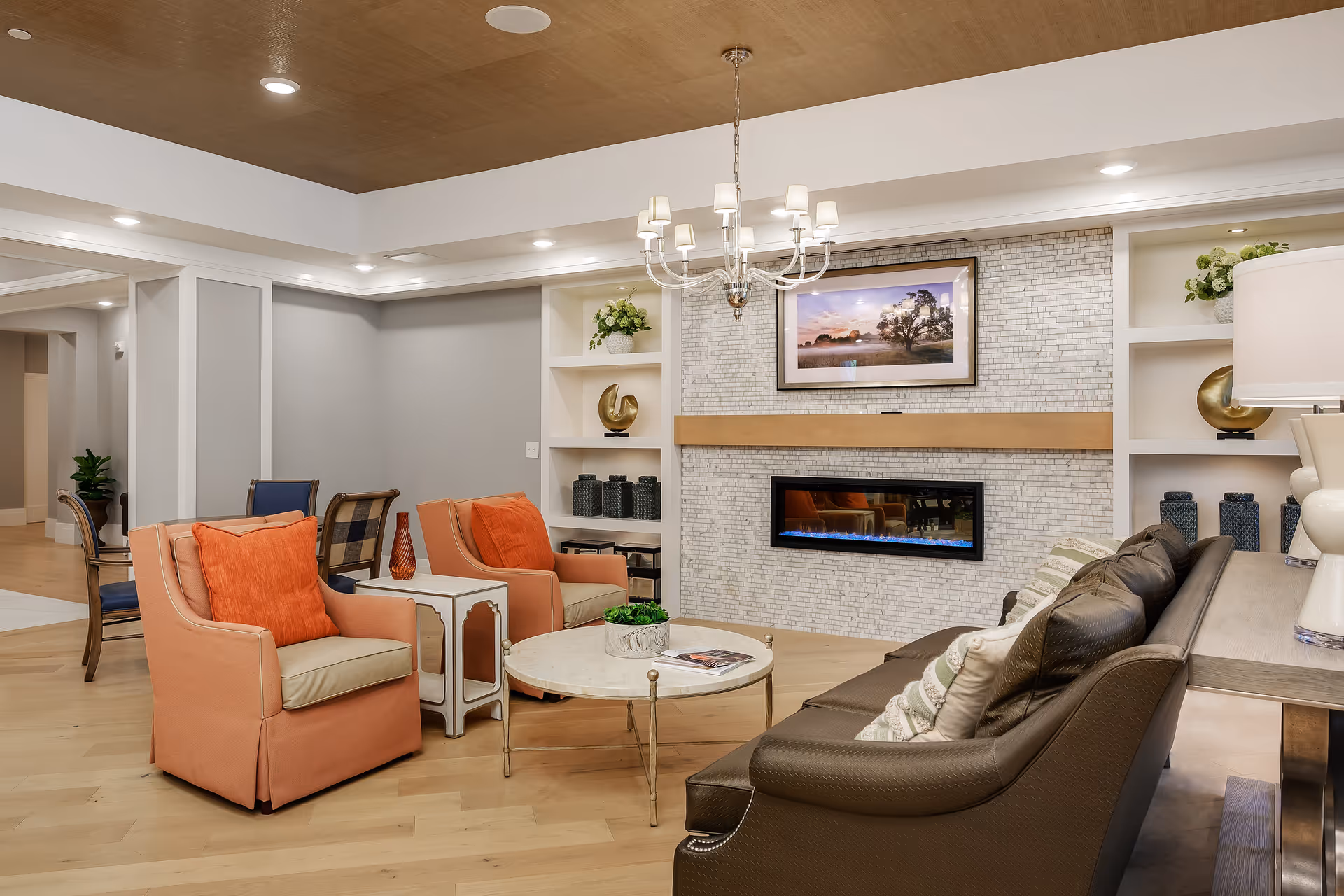A cozy living room area with a modern electric fireplace set in a tiled wall. The room features a brown leather sofa with patterned pillows, two orange armchairs with orange cushions, a round marble coffee table with a small plant and a magazine, and a chandelier hanging from a wooden ceiling. Built-in shelves on either side of the fireplace hold decorative items and plants.