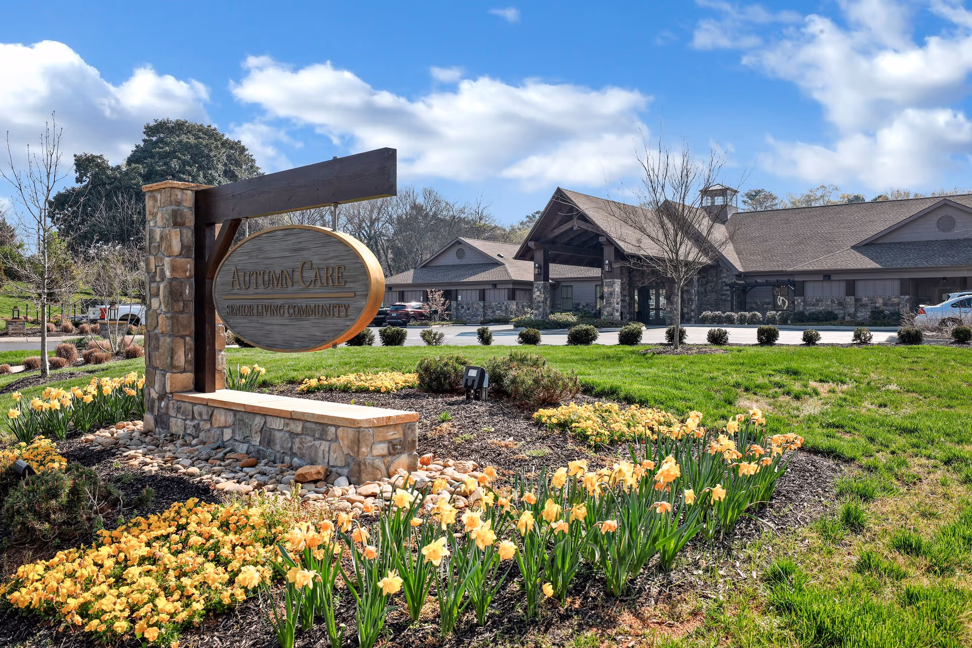 Exterior view of Autumn Care Assisted Living and Autumn Reserve facility with a stone and wood sign in the foreground surrounded by yellow flowers and green grass. The building is visible in the background under a partly cloudy blue sky.