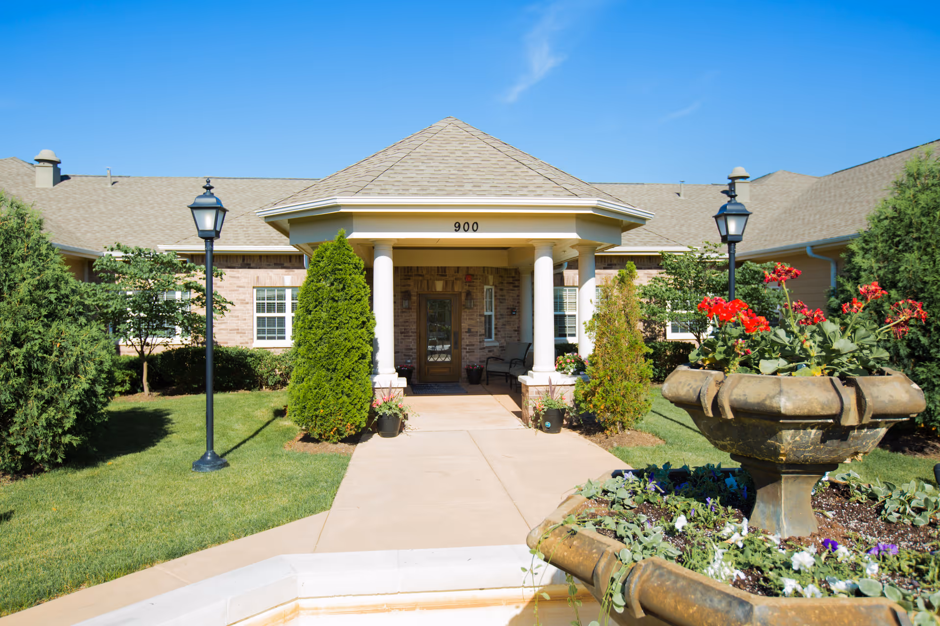 Front entrance of a senior living facility with a covered porch supported by white columns, surrounded by green shrubs and trees. There is a paved walkway leading to the door, flanked by two black lamp posts. In the foreground, there is a stone planter with red and purple flowers. The building has a beige brick exterior and a gray shingled roof under a clear blue sky.