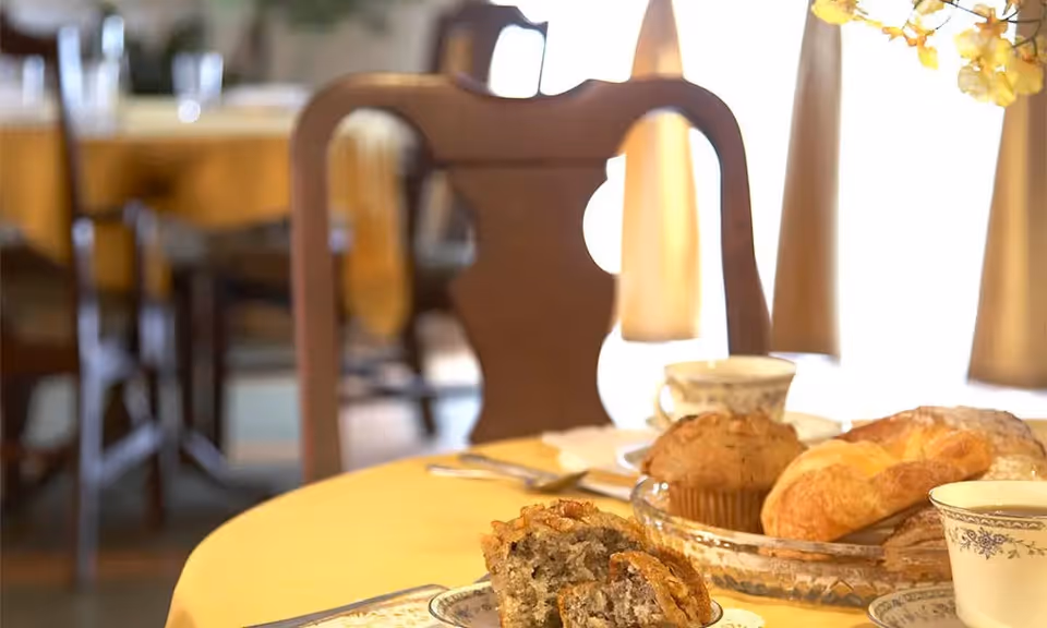 Close-up of a dining table set with a plate of muffins and pastries, fine china cups, and a yellow tablecloth in a warmly lit dining room with wooden chairs and blurred background tables.