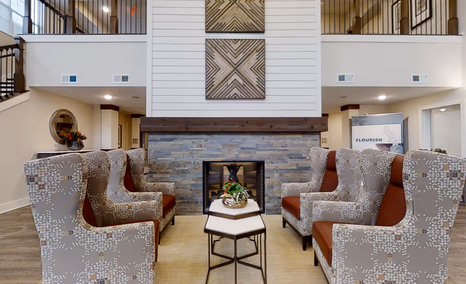 A cozy senior living facility common area with six patterned armchairs arranged around two hexagonal nesting tables. The room features a stone fireplace with a wooden mantle and two geometric wall art pieces above it. There is a staircase with a dark railing on the left and a banner with the word 'FLOURISH' on the right side of the room.