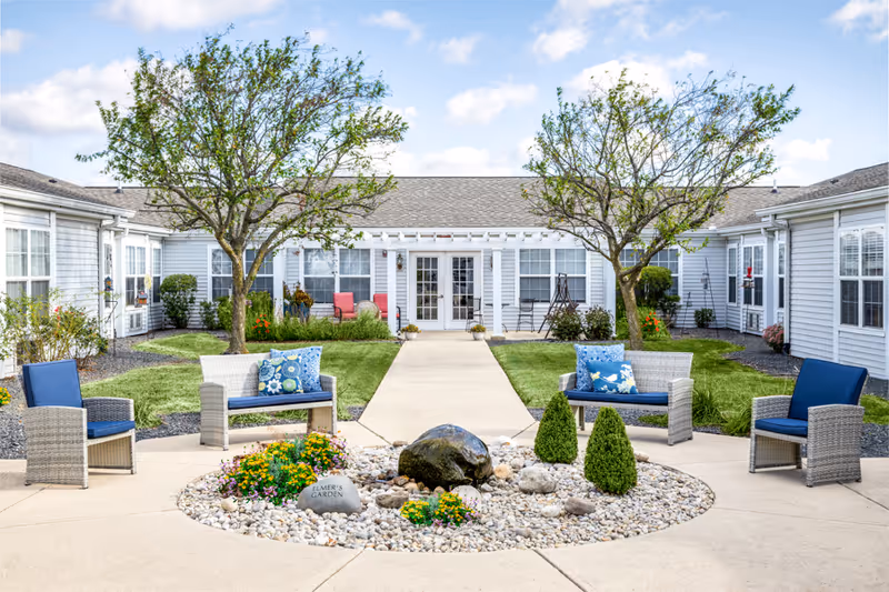Landscaped senior living courtyard with wicker seating around a pebble fountain and a walkway leading to the building's French doors.