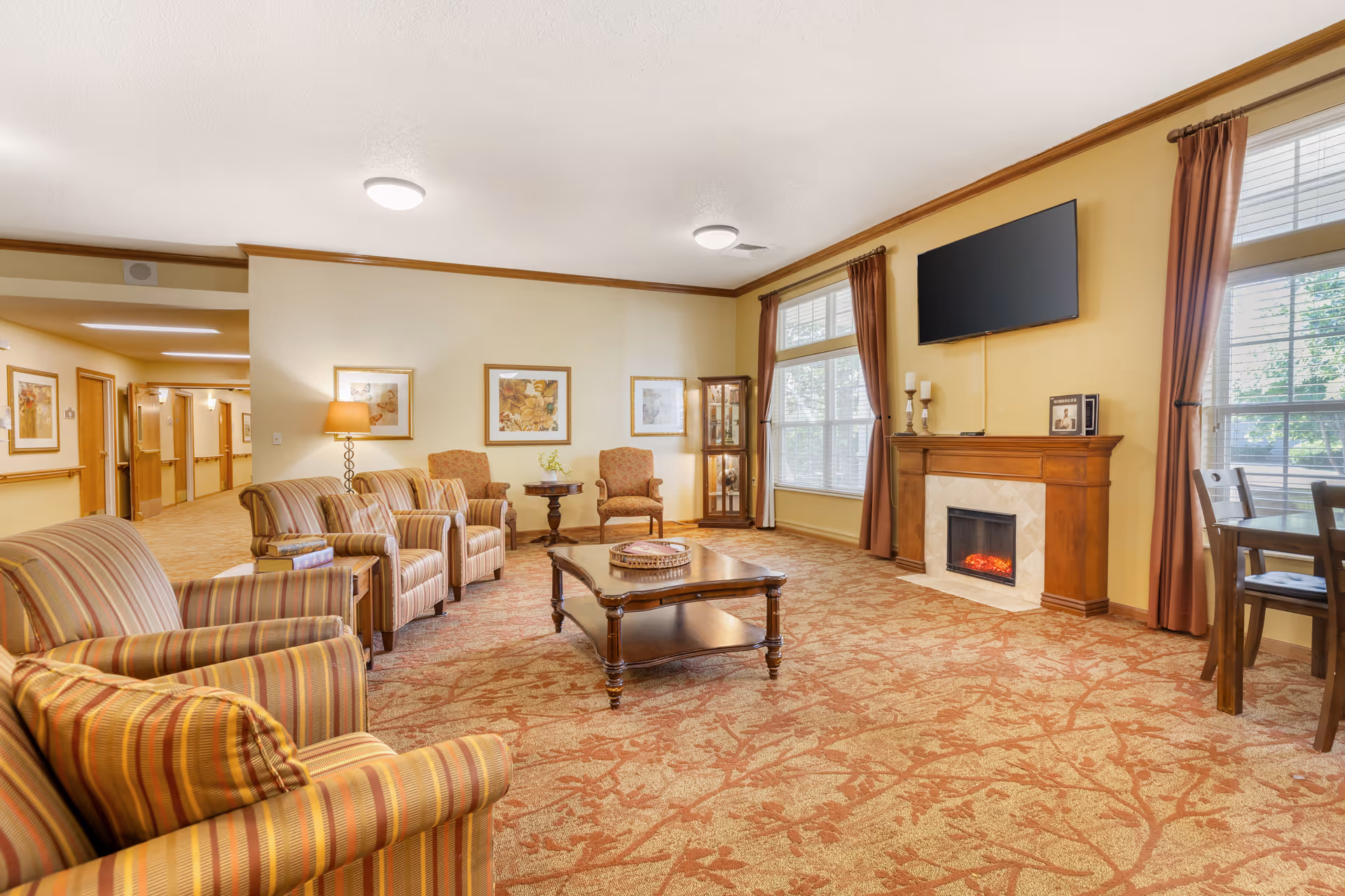 Comfortable senior living common room with striped armchairs, a coffee table, fireplace, wall-mounted TV, and large windows.
