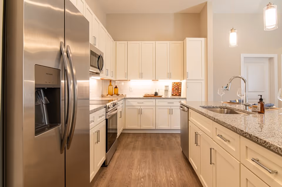 Modern kitchen with white cabinetry, stainless steel refrigerator, oven, microwave, and dishwasher. Granite countertops with a sink and two pendant lights hanging above the island. Wooden flooring and under-cabinet lighting illuminate the space.