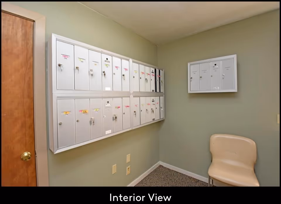 Interior view of a small room with multiple white mailboxes mounted on two walls, a beige chair in the corner, light green walls, a wooden door with a brass knob, and a patterned carpeted floor.